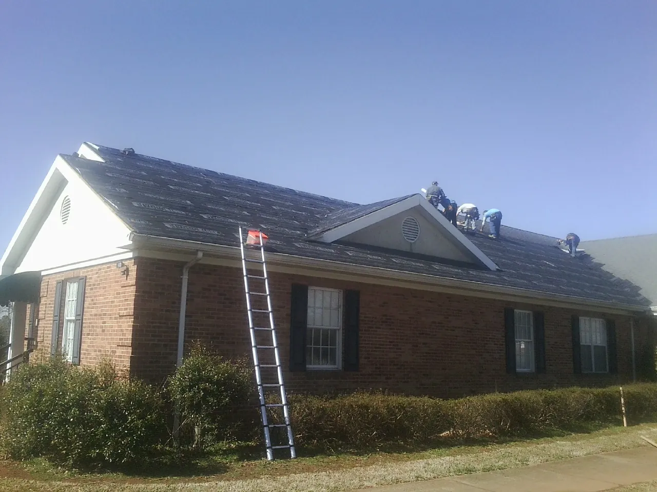 Crew tearing shingles off a brick ranch roof with a ladder set — Emergency Roof Repair work by our crew in Meservey