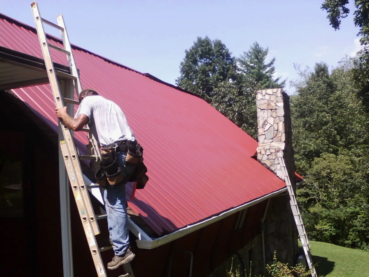 Roofer on a ladder beside a red metal roof and stone chimney — Soffit & Fascia Repair by Roofing Brothers in Meservey